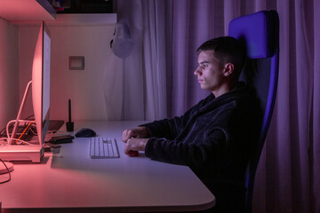 A teenager sits at a desk in a dimly lit room. He focuses on the screen while using a keyboard. The light from the computer illuminates his face during late hours.