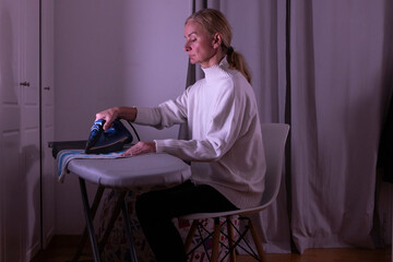 A woman sits at an ironing board in her home. She uses an iron to press a piece of fabric. The room has dim lighting and curtains. It is late in the evening. © Nasos Zovoilis