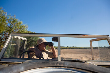 Mature adult male safari guide wearing bucket hat gripping wheel and holding binoculars on savannah