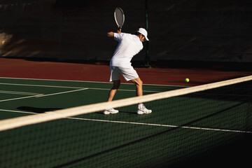 Male tennis player swinging racket at incoming ball on hard court under bright sunlight near net