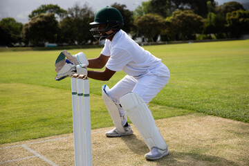 Pacific Islander male athlete crouching behind wickets on pitch wearing white kit and green helmet