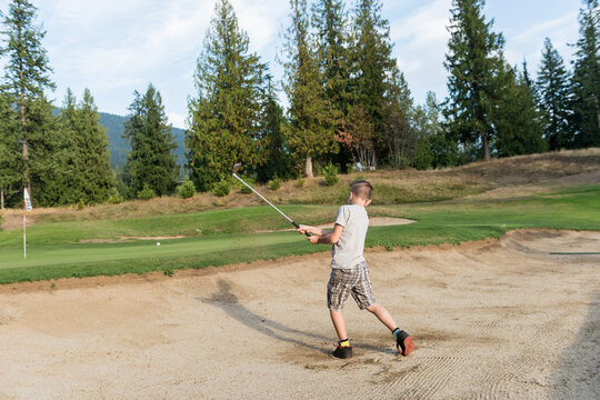 Boy swinging golf club from sand bunker on manicured golf course fairway aiming toward flagstick