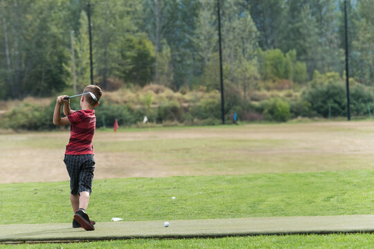 Boy swinging golf club at outdoor driving range near forest on synthetic mat with golf balls