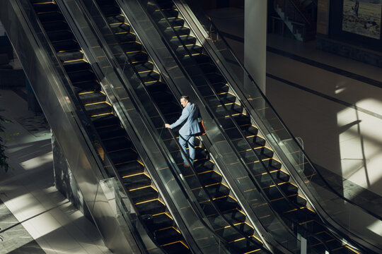 Businessman carrying messenger bag ascending escalator in sunlit lobby past column and wall display