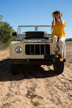 Woman sitting on off-road vehicle fender on track wearing straw hat holding binoculars, copy space