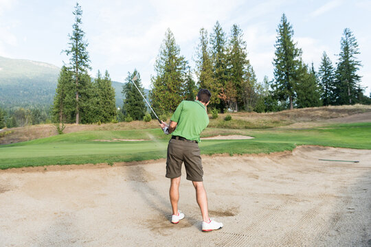 Man swinging sand wedge in bunker toward putting green featuring midair golf ball and metal rake