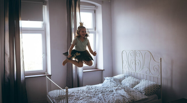 Girl jumping on unmade bed wearing paper crown, tulle skirt with star appliques, copy space