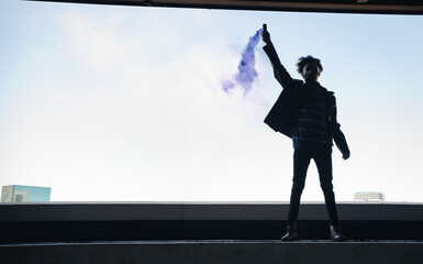 Man holding smoke flare emitting purple smoke on concrete rooftop ledge overlooking skyline