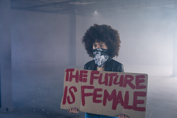 African American adult woman standing in warehouse holding red cardboard sign reading FUTURE FEMALE