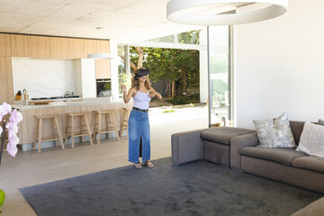 Woman wearing virtual reality headset and holding motion controllers by open-plan kitchen island