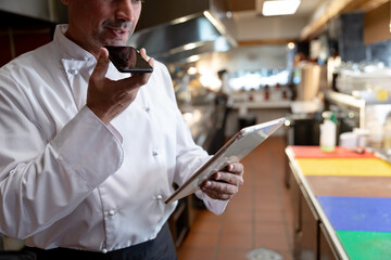 Male chef in uniform using smartphone, tablet at counter with colorful cutting boards in kitchen
