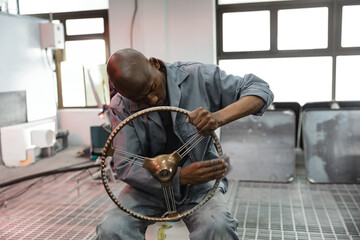 African American male mechanic polishing vintage wheel with wood rim on stool in auto shop