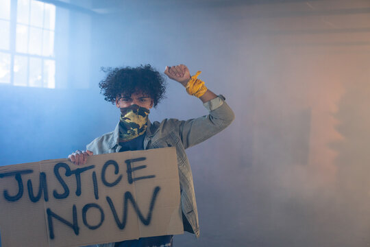 African American man holding protest sign reading JUSTICE NOW in warehouse wearing camo mask
