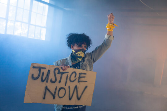 African American man wearing denim jacket raising fist, holding JUSTICE NOW sign in hazy warehouse