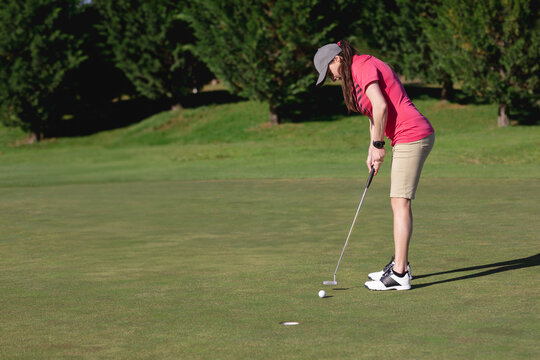 Female golfer in pink polo aligning putter with ball near hole on putting green, copy space