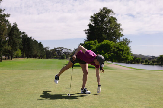 Female golfer gripping putter bending forward placing golf ball on putting green by water hazard