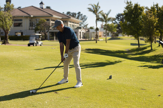 Senior man aligning driver on tee box near clubhouse with golf cart and ball, copy space