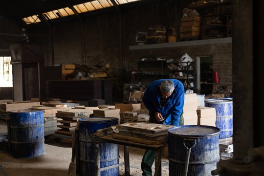 African American craftsman in coverall sanding wood mold on workbench in workshop with blue barrels