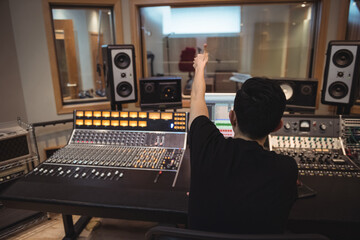 Asian man adjusting faders on mixing console in studio control room with computer screen