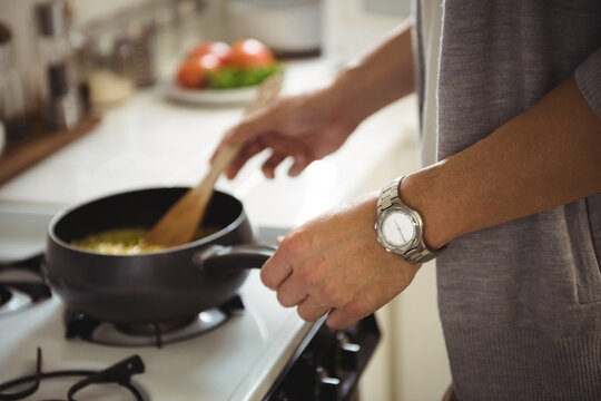 Asian man stirring scrambled eggs in black saucepan on gas burner in home kitchen, copy space