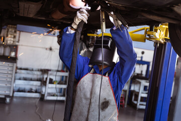 African American female mechanic wearing helmet using torch under lift at workshop, sparks flying