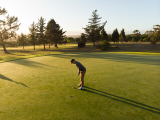 African American man bending over lining up putt on putting green, golf ball near hole, putter