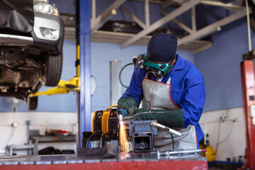 African American woman wearing goggles using grinder over vise sparking near car lift at workshop