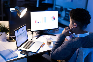 Mid adult African American man sitting at desk under lamp, analyzing data on laptop and monitor