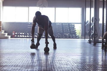 African American man holding kettlebells while performing push-up on gym floor near dumbbell rack