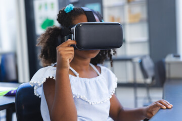 African American girl sitting at desk in classroom wearing virtual reality headset holding front
