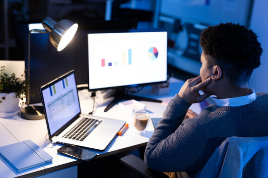 Mid adult African American man sitting at desk under lamp, analyzing data on laptop and monitor