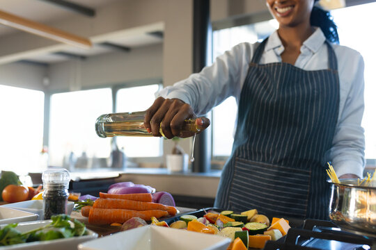 African American woman wearing apron drizzling oil onto vegetables on stove in kitchen, copy space