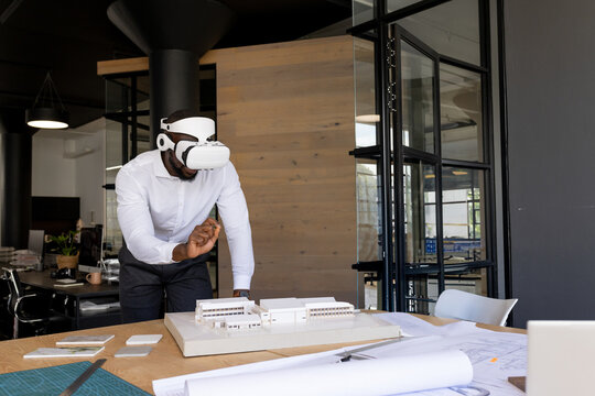 African American man wearing white VR headset, leaning over model and blueprints at office table