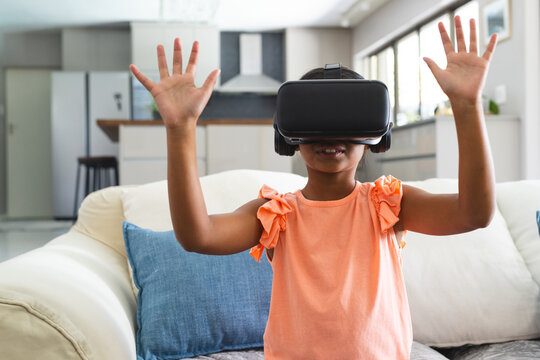 African American girl wearing VR headset playing on sofa near kitchen barstools in open-plan room