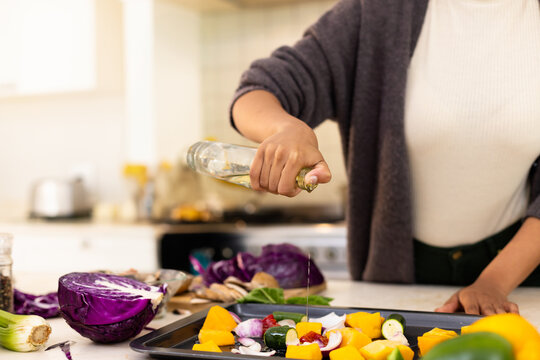 Mid adult African American woman pouring oil from glass bottle onto tray with vegetables in kitchen - Powered by Adobe