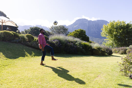 Mid-adult African American man in pink shirt juggling soccer ball on garden lawn by patio umbrella