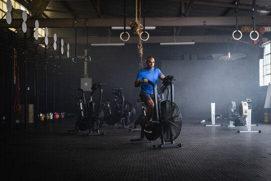 Mid adult African American man riding stationary fan bike at gym wearing sportswear by kettlebells