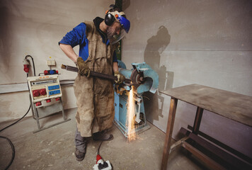Male craftsman wearing safety gear grinding rod at workshop bench grinder sparks flying
