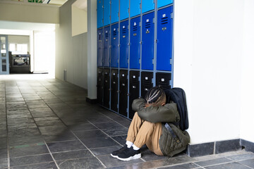 African American boy sitting on school hallway floor hugging knees by lockers, backpack, copy space
