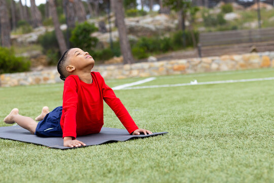 African American boy performing backbend on exercise mat on grass field near stone wall, copy space