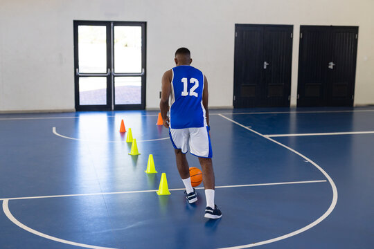 African American male basketball player in blue jersey dribbling orange ball through cones in gym