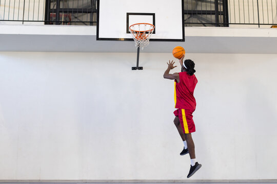 African American male athlete in red jersey dunking at gym hoop holding basketball, copy space