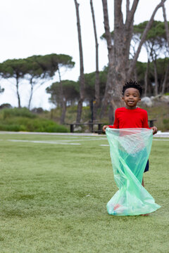 African American boy holding green plastic sack on field in red shirt, blue shorts, copy space