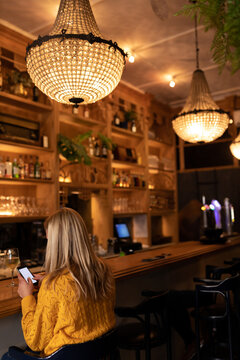 Woman wearing yellow sweater sitting at bar counter using smartphone near wine glass, copy space