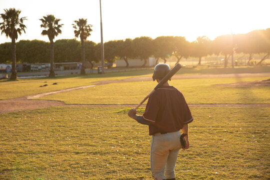 Male baseball player holding bat wearing helmet, glove at sunset with dirt paths and palm trees