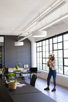 Asian woman standing near office window wearing VR headset using motion controllers, copy space