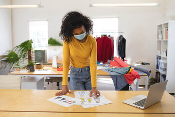 African American woman leaning in fashion studio examining printed sketches and fabric bolts