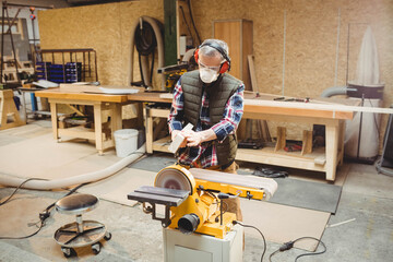 Senior man using disc sander in woodshop wearing safety gear holding wooden block near storage bins