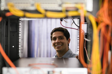 Male network engineer wearing blue shirt smiling inside server rack with yellow ethernet cables