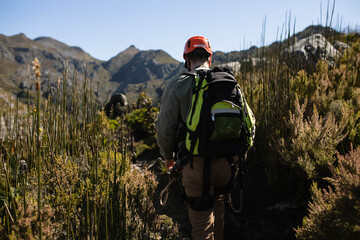 Man in 30s hiking mountain trail wearing red helmet and carrying backpack with rope and carabiners